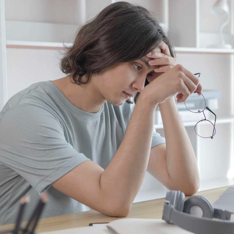 Junge Frau mit langen Haaren sitzt erschöpft am Schreibtisch, hält eine Brille in der Hand und stützt den Kopf nachdenklich ab.