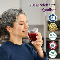 Frau trinkt ein Glas rotes Getränk, entspannt und genießt es. Rechts sind Qualitätszeichen zu sehen: "Ausgezeichnete Qualität", Bio-Qualität, vegan, 36 % mehr Noni-Fruchtanteil, kontrollierte Qualität, sowie ein Siegel für sauberen Sport.